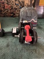 Two black adjustable dumbbell weights with metal handles and a red neoprene dumbbell in front of the bowling bag.