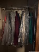 Wide view of men's clothing on hangers, many covered with clear plastic garment bags. Includes shirts, pants, and jackets in various colors such as burgundy and teal.