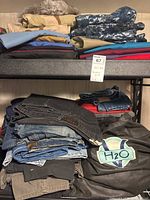 Two shelves with stacks of men's pants including jeans and khakis in multiple colors and styles, with a visible tag and brand logos.