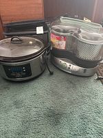 Four kitchen appliances on green carpet: Hamilton Beach slow cooker, Nesco roaster oven, Cuisinart electric fryer, and electric kettle.