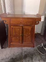 Front view of medium brown wooden cabinet with one drawer and two doors, showing the carved circular motifs and decorative scroll sides.