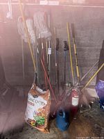 Wide view of garden tools and cleaning equipment against concrete wall showing white mop heads, various hand tools with wooden handles, planting soil bag, watering can, and fire extinguisher.