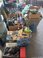 Overall view of lot with various boxes and bins containing hardware, cords, batteries, and office supplies including Milwaukee toolbox visible