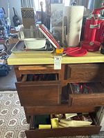 Kitchen countertop with various kitchen items including paper towel holder, red kitchen utensils, metal trays, wrapped coffee pot strainer and drawers below containing more utensils.