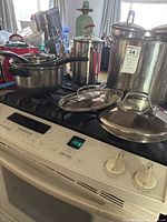 Multiple stainless steel pots and pans with glass lids placed on stove, including large stockpots and saucepans with black handles and red knob lids.