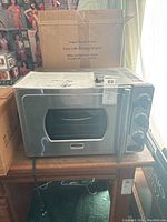 Front and side view of the stainless steel toaster oven with control knobs and instruction manual on top, sitting on a wooden table with boxes behind it.