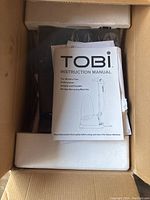 Instruction manual titled 'TOBI INSTRUCTION MANUAL' placed on top of the wrapped steamer inside cardboard box with foam protection.