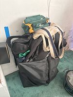 Pile of various tote bags stacked in a black large utility bag on a green carpeted floor against a white wall.