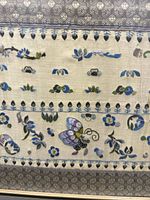 Close-up of the antique Chinese silk embroidery showing detailed butterfly and floral patterns in blue and purple tones on a beige silk background with decorative bordered edges.