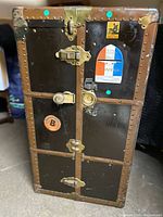 Front external view of weathered black and brown travel steamer trunk with metal rivets and multiple locks, featuring travel labels.