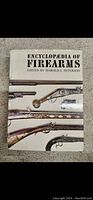 Photo of the front cover of the book 'Encyclopaedia of Firearms' showing historical firearms images and the editor's name.