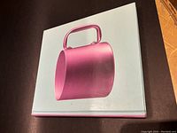 Closed view of the hardcover art book showing the front cover with a pink bucket-like design object on a light blue background.