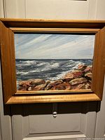 Full view of the framed oil painting showing ocean waves and rocky shore under cloudy sky.