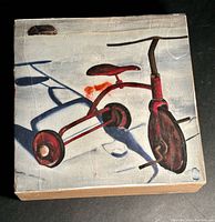 Front view of the original tricycle painting showing the tricycle and its shadow on a light background with red and brown tones on a square wooden block.