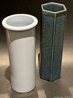 Two vases: a smooth periwinkle blue cylindrical vase and a textured hexagonal blue-green vase standing together on a dark surface.