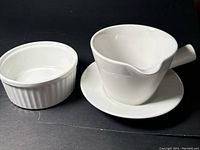 Side view showing white ceramic sauceboat with spout, handle, and matching saucer placed on a flat surface, alongside a small ribbed bowl.