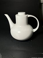 Front view of rounded white porcelain teapot with long spout and handle against a black background