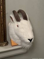 Front view of plush mountain goat head wall mount with white fur, brown horns, and black nose, mounted on wood plaque.