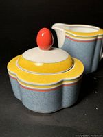 Photo showing lidded sugar bowl with red rounded knob and creamer behind, highlighting postmodern colorful design.