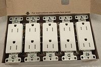 Close-up of five white tamper resistant duplex outlet receptacles still in the original box showing wiring terminals and outlet design.