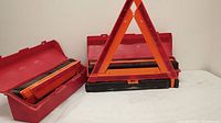 Six orange reflective foldable roadside safety warning triangles with black bases and red carrying cases, some cases open showing folded triangles inside.