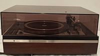 Full view of the Dual 1228 turntable with smoked transparent dust cover closed. Wood grain base and visible control panel.