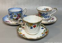 Three vintage teacups displayed with matching saucers, showing floral patterns and gold trim, photographed from the front and slightly above to capture details.