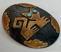 Front view of oval brooch with cat design in gold and turquoise eyes on textured dark silver background.