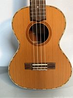 Close-up front view of the ukulele showing solid cedar top, bridge, nylon strings, and decorative binding around the body.