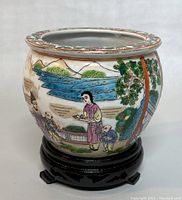 Front view of a rounded ceramic planter showing colorful hand painted Asian figures, water and mountains with floral rim detail, sitting on a black wooden base.