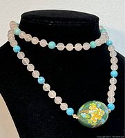 Necklace displayed on a black stand showing the full length with the green cloisonné enamel egg pendant and alternating rose quartz and turquoise beads.