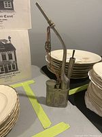 Full view of the antique metal vase shaped like a pipe with floral engravings surrounded by stacks of plates.