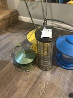 Four bird feeders on the floor: green round with dome and wire base, black cylindrical cage with clear container, blue round, and yellow tray-like feeder