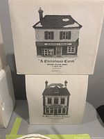 Front of boxes featuring illustrations and names of the two porcelain village houses from Department 56 Dickens Village Series.