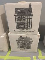 Boxes of two Department 56 collector ceramic buildings stacked, labeled 'Shops of Dickens Village' and 'Dickens Lane'