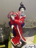 Full view of Geisha statue showing red kimono with floral patterns, holding a fan, standing on a base.