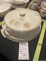 Full shot of the soup terrine with lid atop a serving platter, cream-colored ceramic with brown and gold leaf floral design and gold accents around edges and lid knob.