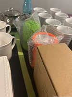 Two plastic collapsible lanterns wrapped in bubble wrap, one orange and one bright green, with packaging box labeled Collinsible Lantern Set. Surrounding white cups visible.