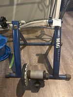 Front view of blue MAG manual bike trainer showing frame and resistance roller unit on floor.