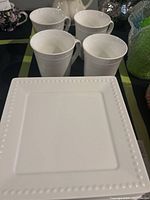 Four white ceramic mugs and one white square plate with bubble dot border pattern on a dark surface