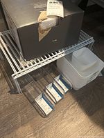 White metal shelf with boxed item on top, white plastic containers to side, and four clear plastic packet organizers underneath