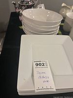 Stack of eight white square plates with a MaxSold tag indicating 'Square dishes + misc'. Plates show some chips and need cleaning.