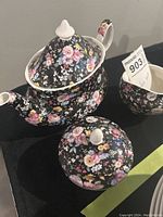 Photo showing whole tea set: teapot with lid, sugar bowl with lid, and creamer with black base and dense multicolor floral pattern