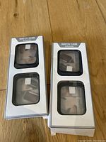 Two unopened white cardboard packages with transparent front windows showing two black Apple Watch protective covers each inside, placed on wooden background.