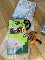 Two soft fabric baby books titled 'Jungly Tails' with colorful jungle animal designs and packaged in clear plastic bags, one book outside the bag showing a bright green cover with a monkey and elephant.