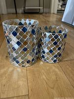 Front view of two blue tone metal candle holders with diamond lattice pattern placed on a wood floor.
