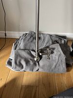 Grey microfibre curtains folded with a silver metal curtain rod and mounting bracket placed on top on a wood floor in front of a beige wall.