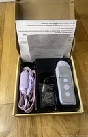 Open box view showing electronic dog collar with lavender strap, remote control with three buttons, charging accessory, and instruction manual.