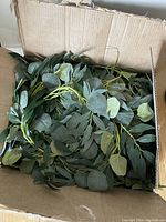 Photo of two large faux eucalyptus and willow leaf garlands loosely coiled inside a cardboard box.