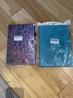 Two tablet/iPad cases in packaging, one purple mandala pattern and one teal with embossed floral design.